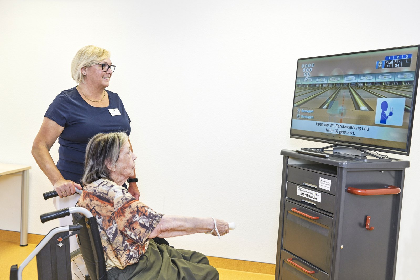 Patientin im Rollstuhl spielt Wii bowling unter Aufsicht einer Pflegeperson